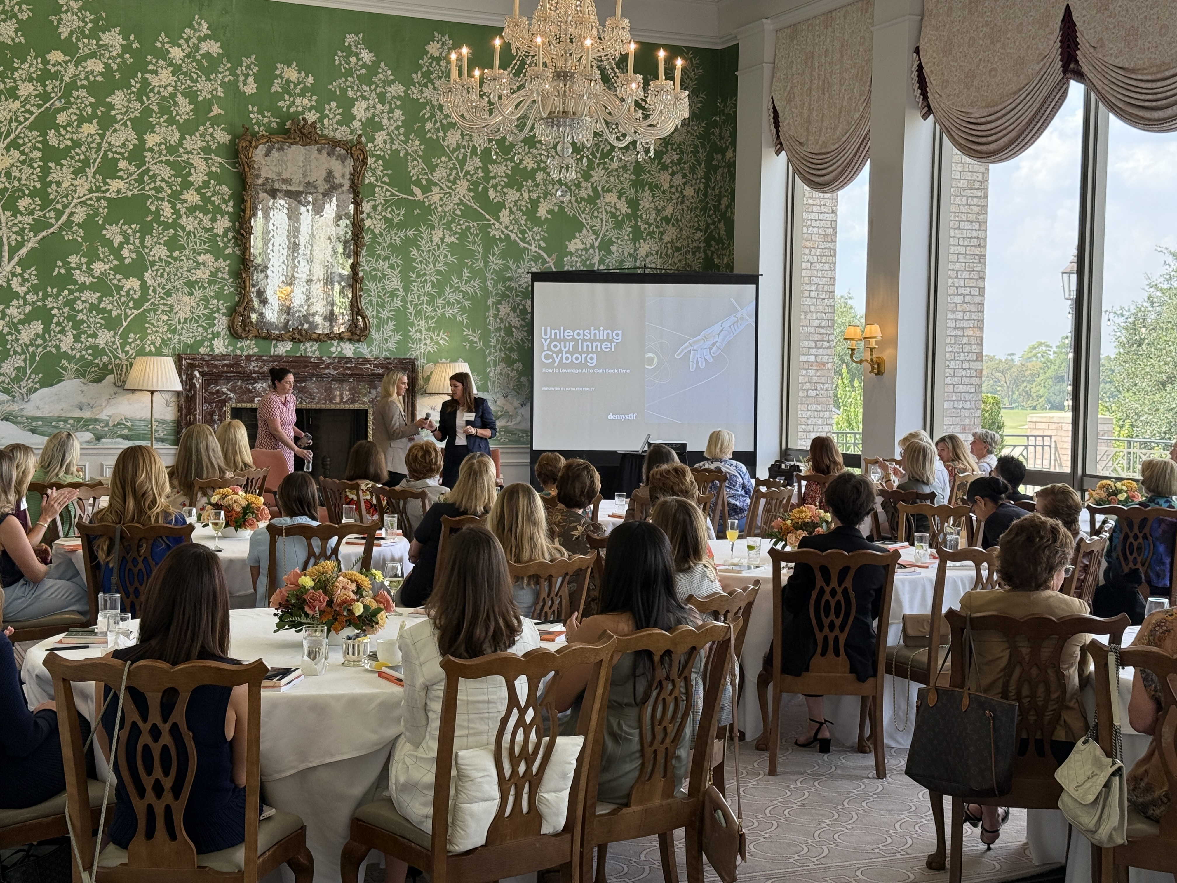 Speaker presenting in room with tall ceilings and a medium screen for presentations, floor to ceiling windows on one wall, floral green wallpaper on the other, spectators seated at round tables