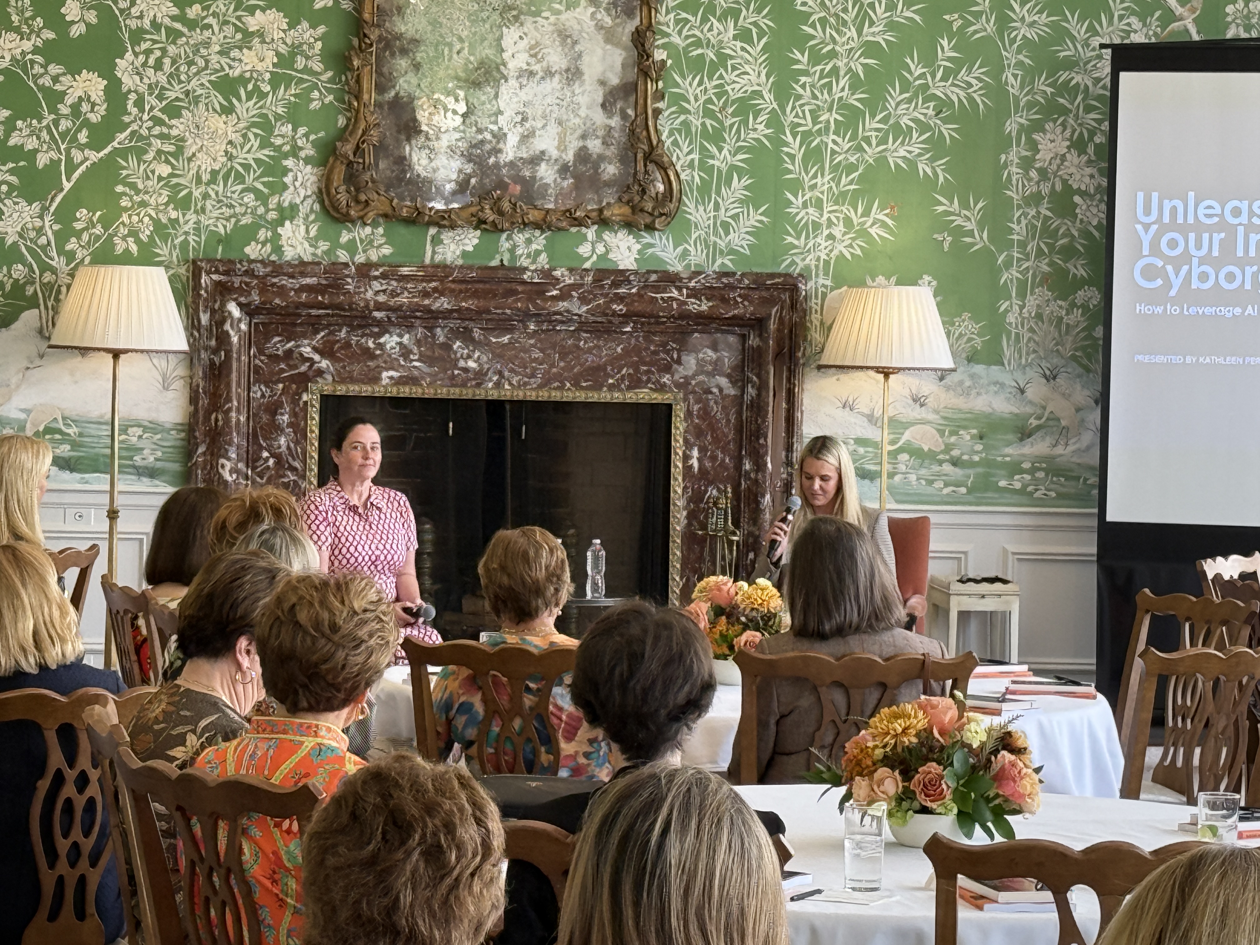 Speaker presenting in front of a dark marble fireplace, spectators seated at round tables